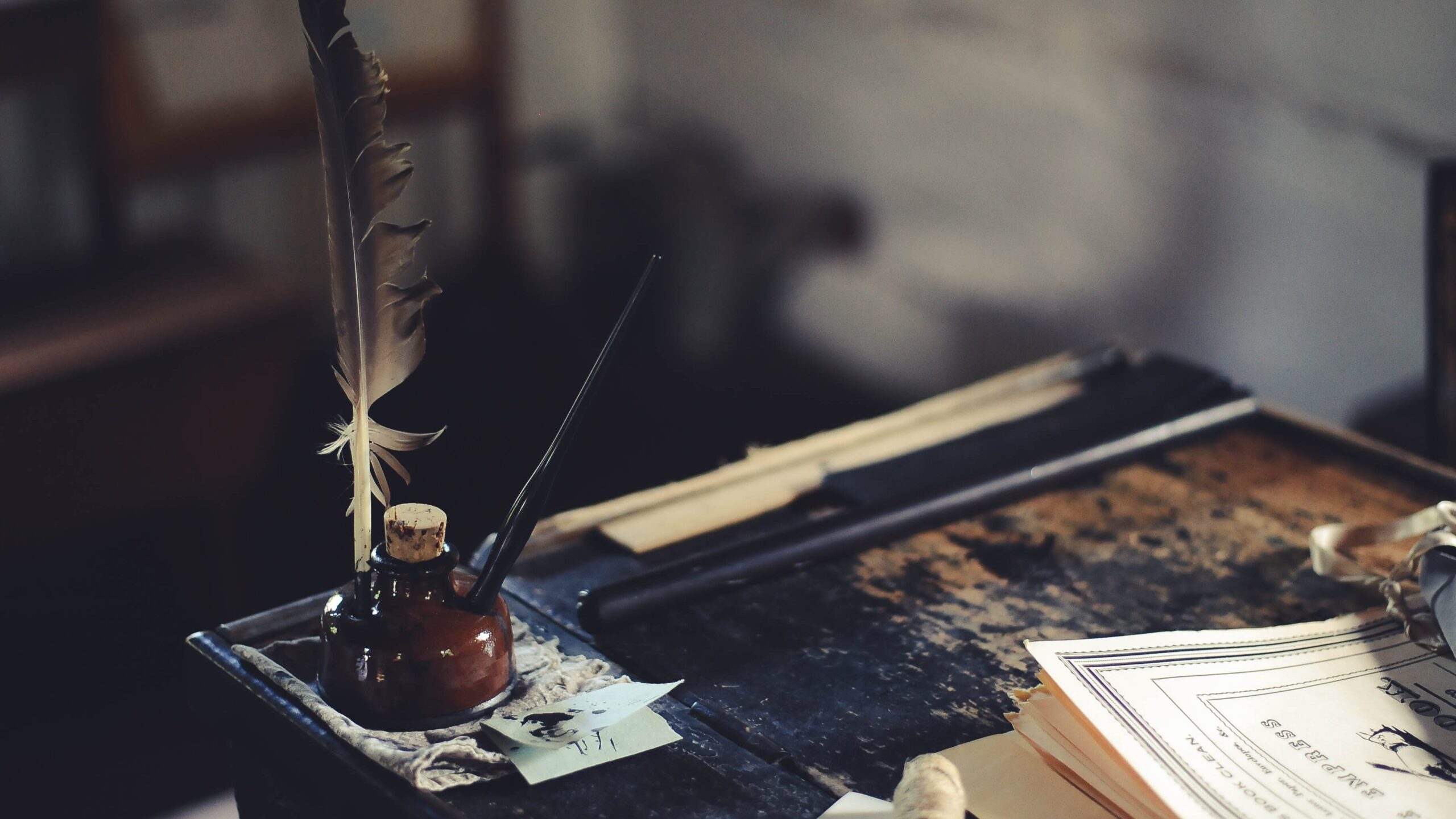 A bottle of ink with a quill on an old wooden desk with papers and books to the side 