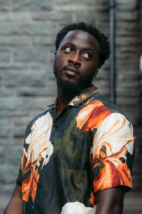 Author Nana Kwame Adjei-Brenyah, a Black man in a colorful shirt against a gray brick background.