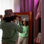 A Black teen girl in a green top tries on a spotted hat in front of a mirror