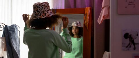 A Black teen girl in a green top tries on a spotted hat in front of a mirror