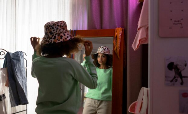 A Black teen girl in a green top tries on a spotted hat in front of a mirror