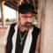 Bearded man in glasses and black cap wearing a white shirt and black vest and undershirt leaning against a wall and looking to the viewer's right.