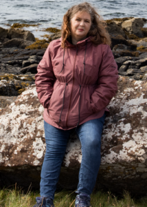 Author Melissa Marr, a woman in a jacket sitting on a rock by the shore