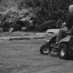 A black-and-white photo of a bald man riding a lawnmower over the grass of a lawn.