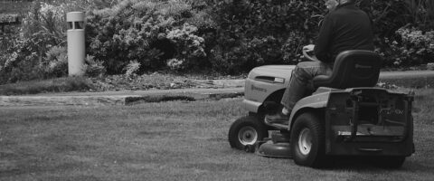 A black-and-white photo of a bald man riding a lawnmower over the grass of a lawn.