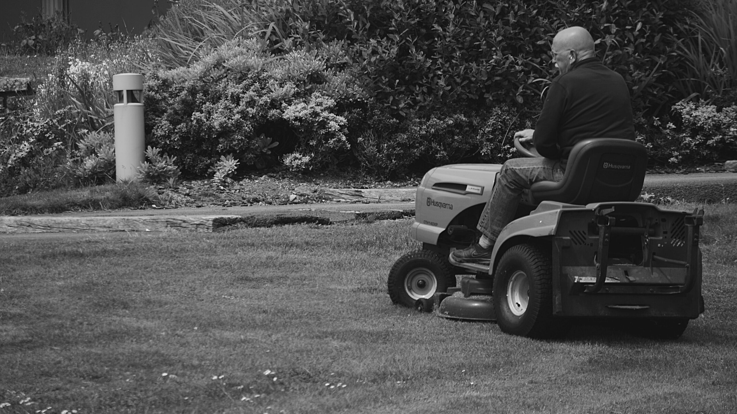 A black-and-white photo of a bald man riding a lawnmower over the grass of a lawn. 