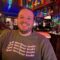 Smiling bearded man wearing a sweatshirt that says "Read More Queer Books" in a bar with LGBTQ+ rainbox flags hanging from ceiling .