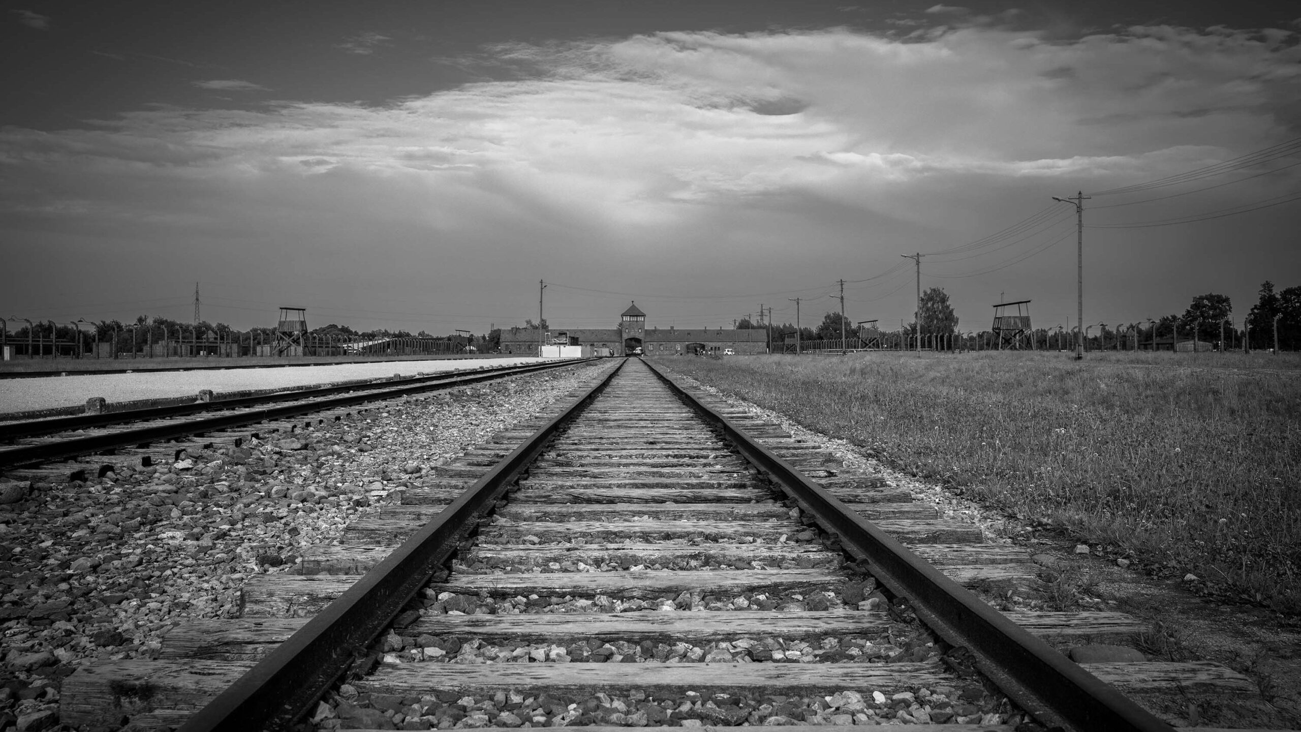 A black-and-white photo showing train tracks leading to the entry for Auschwitz under a cloudy sky through which the sun's glow can be seen.