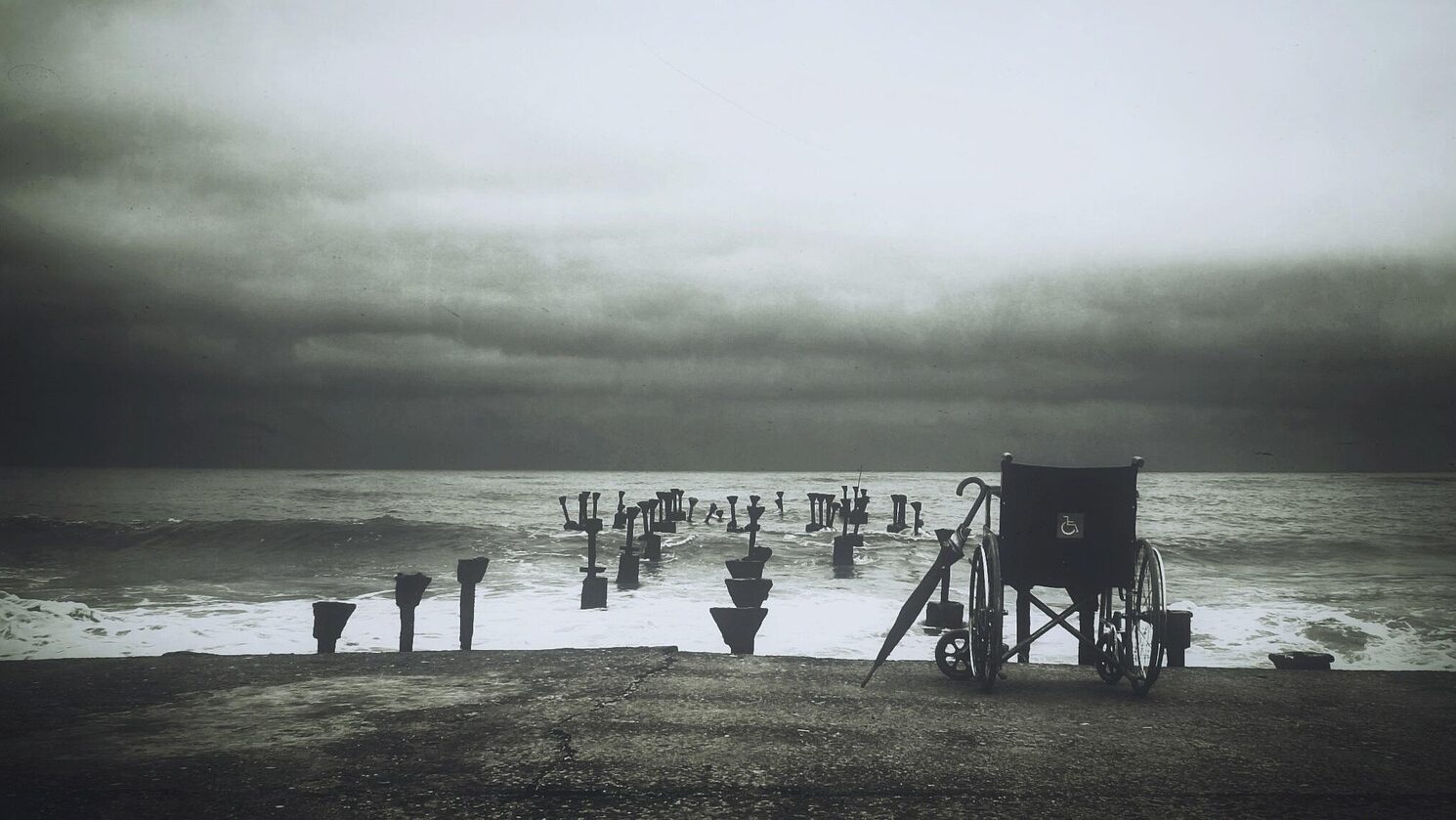 A black-and-white photo of wheelchair and umbrella sitting abandoned before the ruins of an old pier that extends out into the ocean.