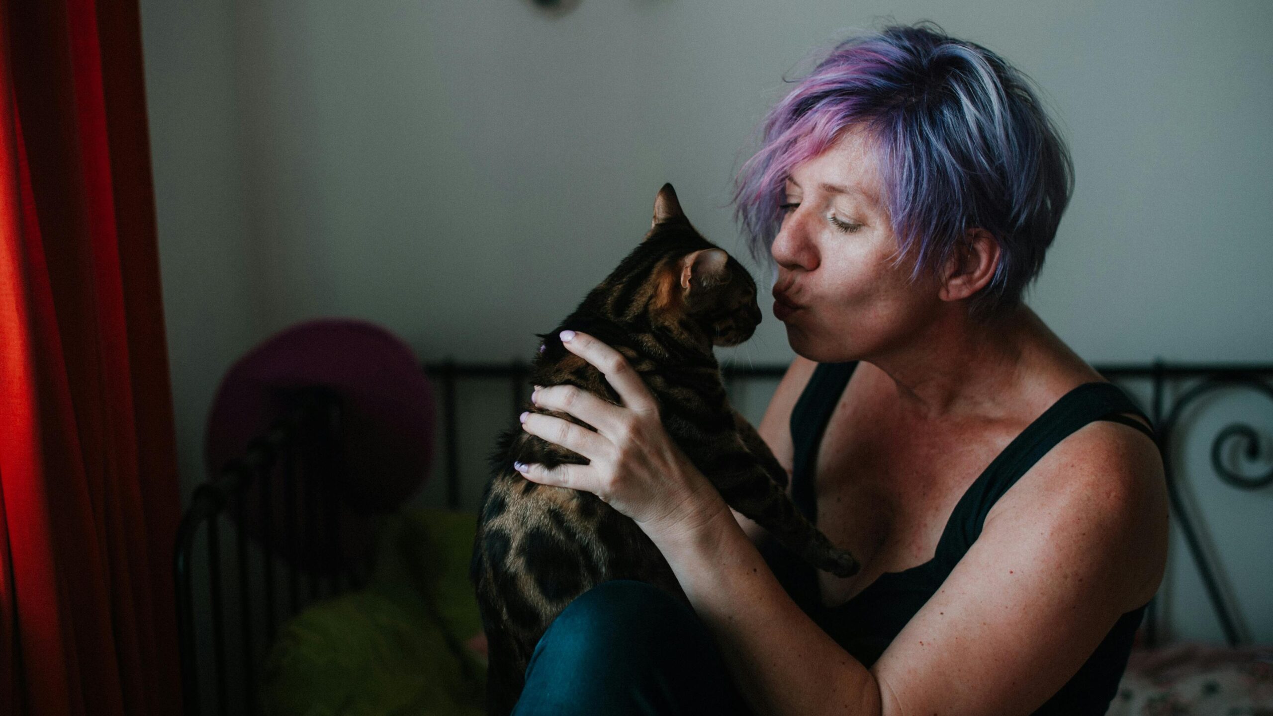 A middle-aged white woman with purple-streaked hair and a black tank top holding up a tabby cat and kissing it on the nose.