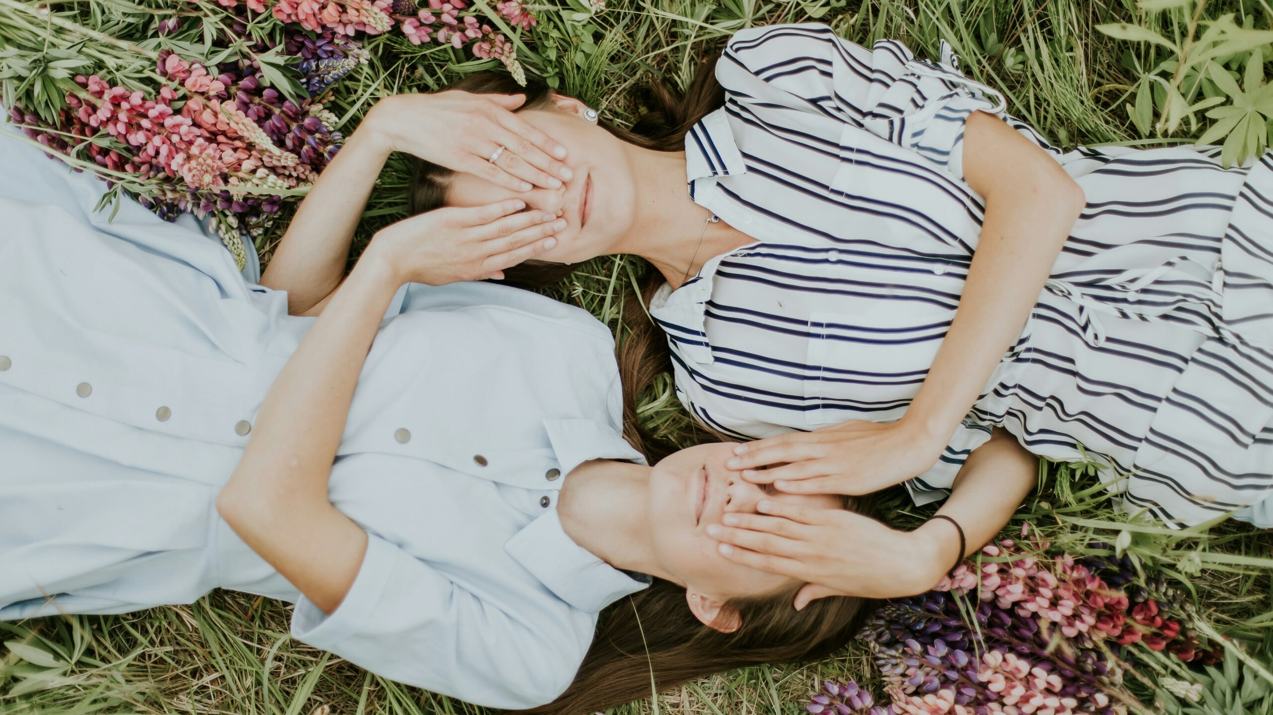 Two sisters lying down in a field in opposite directions, face up, one in a plain light blue dress, the other in a white dress with black stripes, with each others hands covering the other sister's eyes.