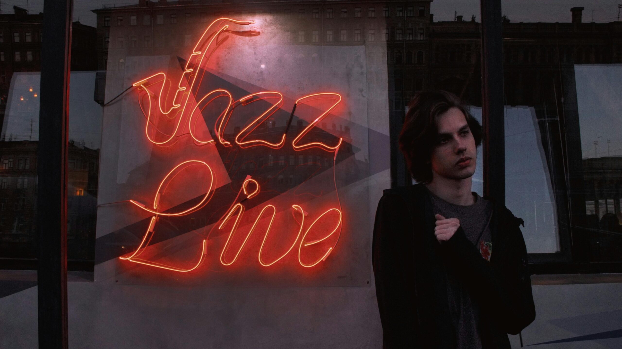 A pensive dark-haired white man beside a neon sign for Live Jazz.
