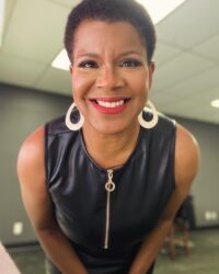 Photo of Tananarive Due, a smiling Black woman with big gold hoop earrings in a leather dress leaning toward the camera and smiling.