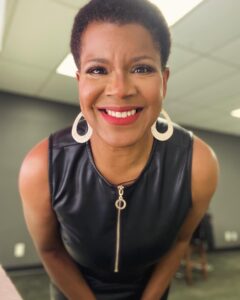 Photo of Tananarive Due, a smiling Black woman with big gold hoop earrings in a leather dress leaning toward the camera and smiling.