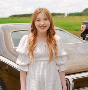 Ashleigh Worley, a smiling white woman with long blond hair who is wearing a white dress and standing in front of a brown Camaro parked beside a field.