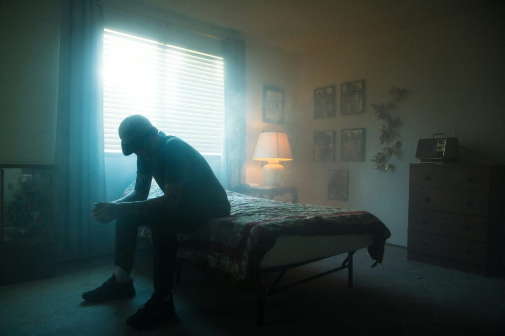 In a dark room, the silhouette of a man sits on a bed, elbows resting on his knees, bowed forward. Behind him, sunlight glows through drawn drapes and a lamp shines dully.