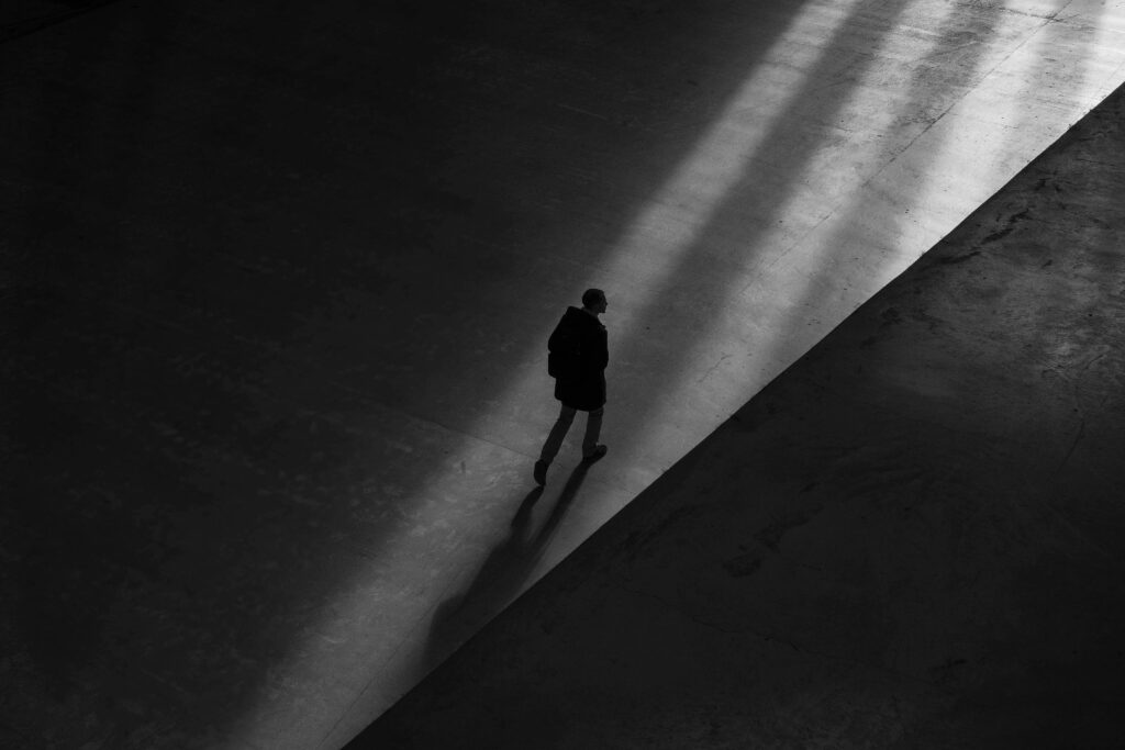 Black-and-white photo with the silhouette of a lone man walking into a beam of light with shadows stretching toward him and his own shadow stretching behind.