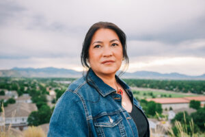 Asian-American woman with black hair and a blue jean jacket looking up outside with a background of gray clouds and mountains
